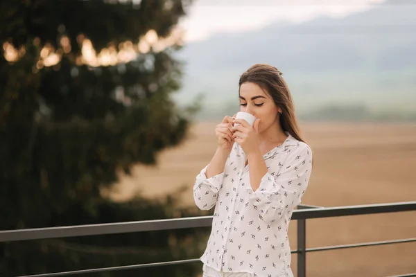 Fiatal lány kávét iszik a teraszon. Háttér, a hegyek és a fenyő. Tavaszi — Stock Fotó