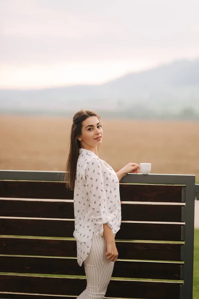 A szép lány a fehér ruhát, kávét iszik a teraszon. Háttér-hegység — Stock Fotó