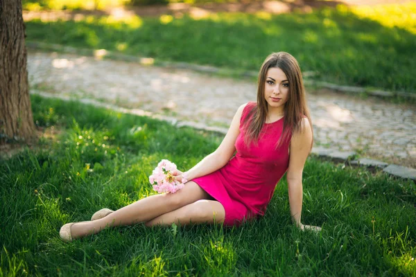 Niña sentada en la hierba y sosteniendo una flor de sakura en sus manos. Antecedentes del bokeh rosa de los árboles de sakura —  Fotos de Stock