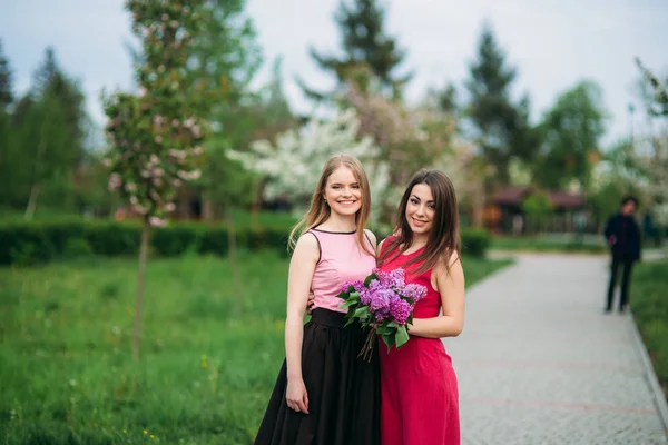 Zwei Schwestern, die draußen im Frühlingspark spazieren. sie halten einen Strauß Flieder und lächeln — Stockfoto