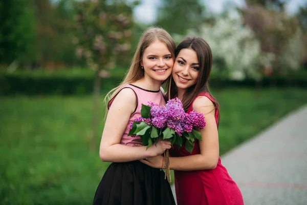 Dos hermanas caminando en el parque de primavera. Llevan un ramillete de lila y sonríen —  Fotos de Stock