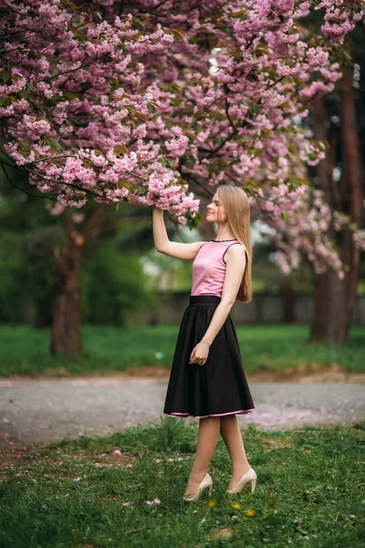 Muchacha atractiva en pie vestido en el parque junto al árbol de flores. Cabello rubio femenino —  Fotos de Stock