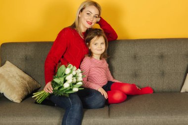 Anne ve kızı kanepe evde oturur ve eğlenin. Beyaz Lale bir buket var. Güzel çiçekler. Sarı arka plan. Ev konfor
