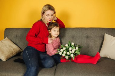 Genç anne gri koltukta oturan kızı ile. Studio sarı arka plan. 8 Mart çiçekler