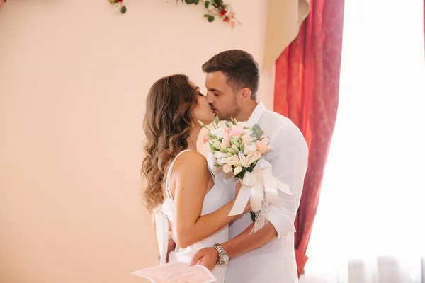 Φιλί του όμορφο ζευγάρι. Γαμπρός στο λευκό πουκάμισο φιλί του κομψή νύφη σε λευκό φόρεμα. Μπουκέτο με λουλούδια στα χέρια — Φωτογραφία Αρχείου
