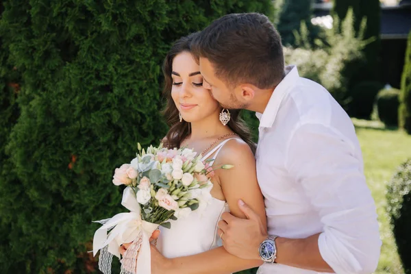 Um homem barbudo e bonito beija a mulher. Mulher gogeous em vestido branco com belo buquê. Homem elegante em camisa branca — Fotografia de Stock