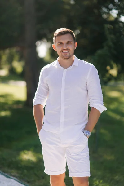 Un hombre guapo con una camisa blanca y pantalones cortos sonríe. Modelo masculino afuera. Fondo del parque. Joven hombre atractivo caminando en el parque — Foto de Stock