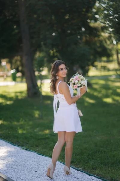 Sexy vrouw in prachtige witte jurk met boeket bloemen wandelen in het park. Vrouwelijke model buiten — Stockfoto