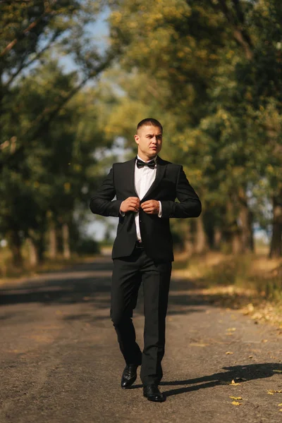 Fiatal vőlegény öltöny otside-ban. Jóképű férfi fekete öltöny. Modell séta a parkban — Stock Fotó