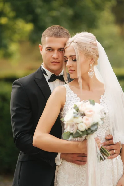 Guapo novio abrazar a su hermosa novia. Recién casados caminando por el parque. Mujer pelo rubio en vestido de novia elegante — Foto de Stock