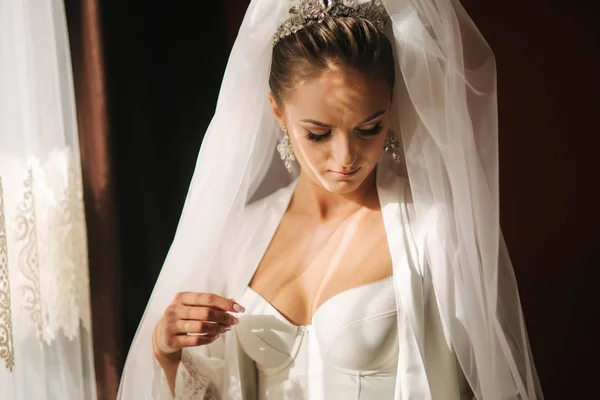 Retrato de noiva bonita em casa. Mulher ficar ao lado da janela no vestido de noiva . — Fotografia de Stock