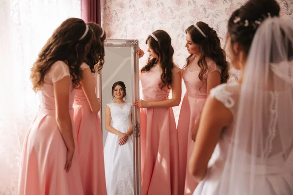 Damigella d'onore in piedi accanto allo specchio e aiutare la sposa a guardare te stesso. Grande specchio a casa. Ragazze felici con lo stesso vestito — Foto Stock