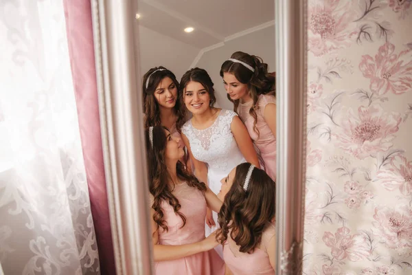 Bridesmaid standing by the mirror and help bride look at yourself. Big mirror at home. Happy girls in same dress