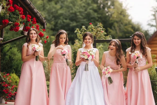 Felice sposa con damigella d'onore tenere mazzi di fiori e divertirsi fuori. Bella damigella d'onore in abiti stessi stand dalla sposa affascinante in abito da sposa lungo — Foto Stock