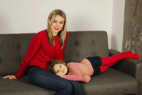 Mama blond păr în pulover roșu așezat pe canapea cu fiica ei. Frumoasa femeie — Fotografie, imagine de stoc