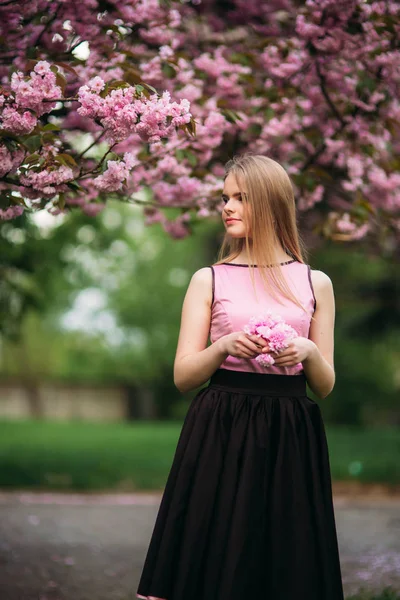 Attraente ragazza in abito stand in parco vicino all'albero dei fiori. Capelli biondi femminili. Sakura di albero rosa — Foto Stock