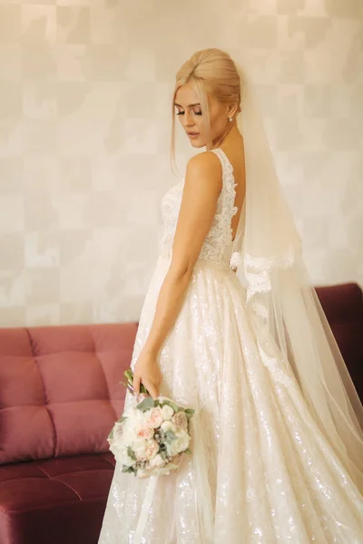 Элегантная невеста в красивом свадебном платье с букетом дома. Блондинка с красивым макияжем и прической — стоковое фото