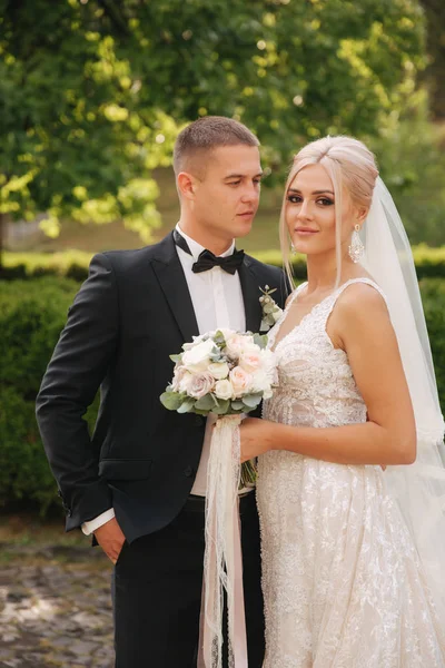 Guapo novio abrazar a su hermosa novia. Recién casados caminando por el parque. Mujer pelo rubio en vestido de novia elegante — Foto de Stock