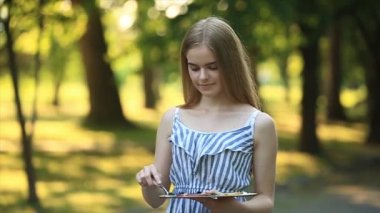 Taburede oturan güzel sarışın kız ve parkta boyalar ve bir spatula ile bir palet kullanarak bir resim çizer