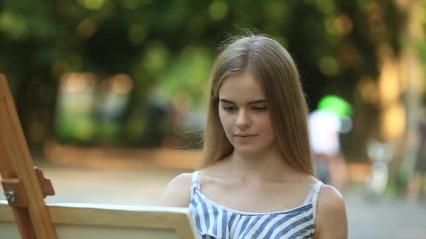 Όμορφο κορίτσι τραβάει μια εικόνα στο πάρκο χρησιμοποιώντας μια παλέτα με χρώματα και μια σπάτουλα. Καβαλέτο και καμβάς με την εικόνα — Αρχείο Βίντεο