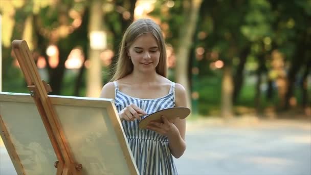 Menina bonita desenha um quadro no parque usando uma paleta com tintas e uma espátula. Easel e lona com uma imagem — Vídeo de Stock