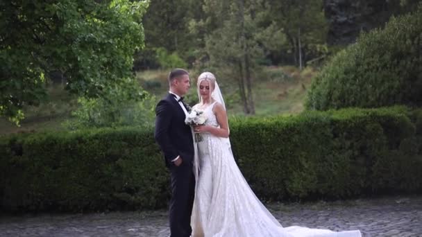 Schönes Hochzeitspaar beim Spazierengehen im Park. Braut mit langem weißen Kleid und stilvollem Bräutigam — Stockvideo