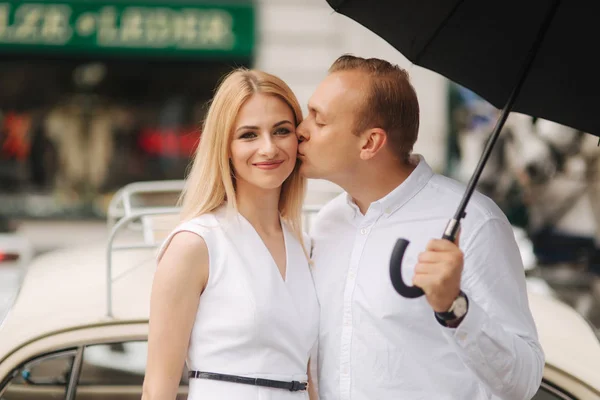 Man and woman stand near old car with umbrella. Beautiful couple in the city. Black umbrella. Black and white clothes