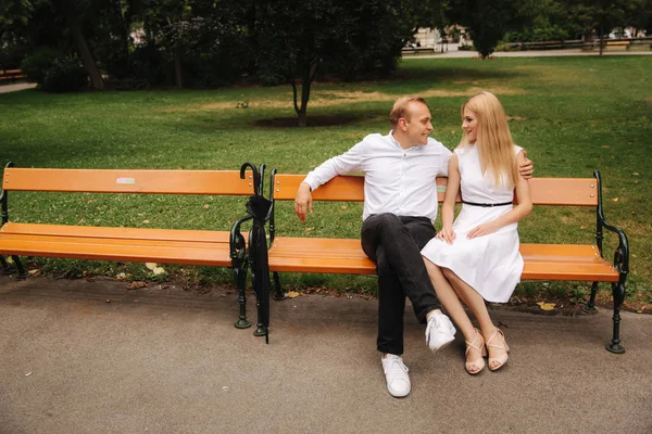 Mooie paar zit op de Bank in het park. Blonde haren man en vrouw besteden tijd togerther. Zij zwarte paraplu. Achtergrond van groen gras — Stockfoto