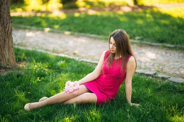 Niña sentada en la hierba y sosteniendo una flor de sakura en sus manos. Antecedentes del bokeh rosa de los árboles de sakura —  Fotos de Stock