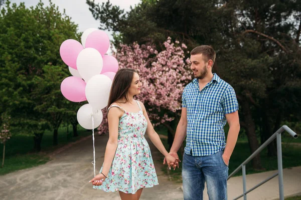 Молода щаслива пара закохана на відкритому повітрі. Красивий чоловік і красива жінка на прогулянці в парку весняного цвітіння. Вони тримають гелієві кульки — стокове фото