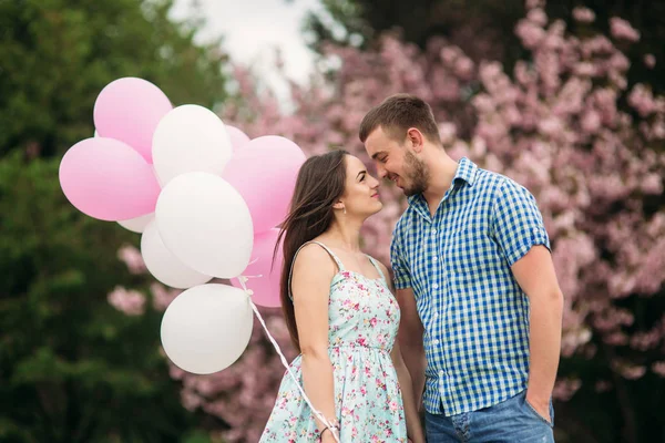 Mladý šťastný pár v lásce venku. Pohledný muž a krásnou ženou na procházce v parku kvetoucí na jaře. Udržují helium koule — Stock fotografie