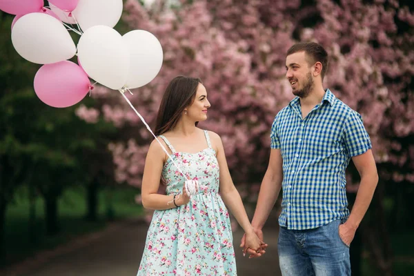 Mladý šťastný pár v lásce venku. Pohledný muž a krásnou ženou na procházce v parku kvetoucí na jaře. Udržují helium koule — Stock fotografie