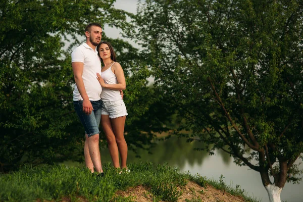 Mladý pár v bílém stojí na kopci. Pozadí stromů a jezero — Stock fotografie