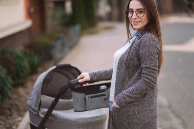 Aile, çocuk ve ebeveyn konsepti-parkta puset ile mutlu anne. Güneş gözlüğü ve şık gri ceket ile anne