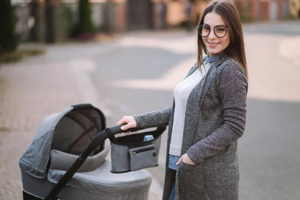 Aile, çocuk ve ebeveyn konsepti-parkta puset ile mutlu anne. Güneş gözlüğü ve şık gri ceket ile anne