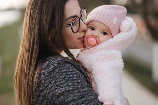 Portré az anya és az ő kis baba lány. Szép anya és aranyos baba. Anya HUD lányát. Baby öltözött peanch színes kalap és a mellbimbó játék. Anya megcsókolja a lányát — Stock Fotó