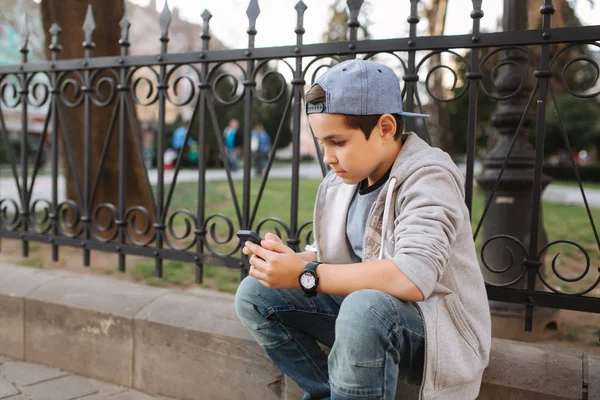 Мальчик играет в онлайн игры на смартфоне снаружи. Мальчик сидит в центре города. Мальчик играет в игры, пока его мама идет в магазин — стоковое фото