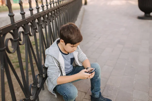 Молодий хлопчик грає в онлайн гру по смартфонах на вулиці. Хлопчик сидить у центрі міста. Хлопчик грає в ігри, поки його мама йде в магазин — стокове фото
