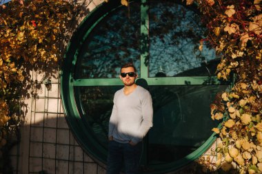 Güneş gözlüğü şık adam yuvarlak pencerenin önünde stand. Sonbahar hava, sarı ve kırmızı yaprakları