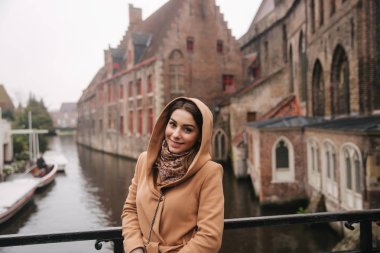 Bealgium, Brugge köprüsünde kahverengi ceket ayakta güzel kadın portresi. Kadın bir Hood koymak