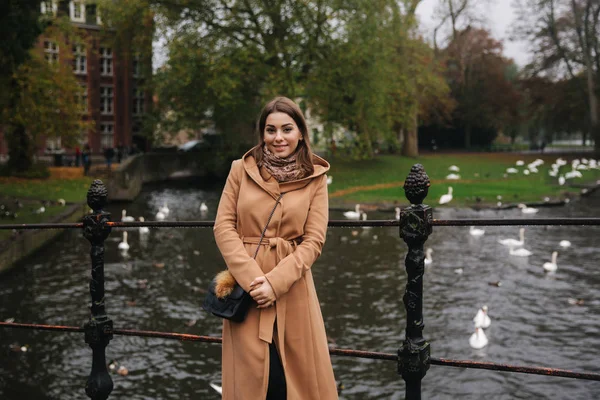 Привлекательная женщина в коричневом пальто, стоящая на мосту, на фоне реки с лебедями — стоковое фото