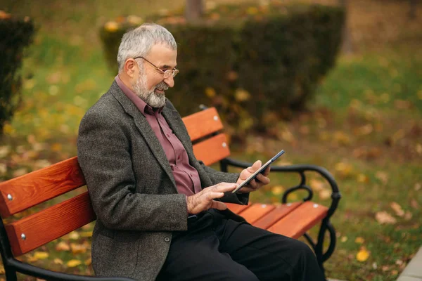Ο παππούς χρησιμοποιεί ένα δισκίο που κάθεται στο πάρκο στον πάγκο — Φωτογραφία Αρχείου