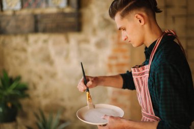 Young artist making ornament on ceramic plate. Handsome young man working at his workshop