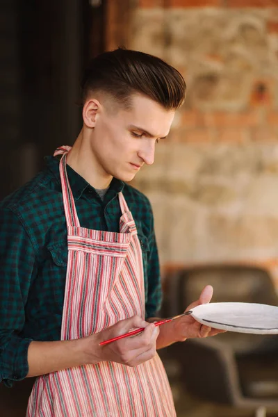 Jonge kunstenaar maken ornament op keramische plaat. Knappe jongeman aan het werk in zijn werkplaats — Stockfoto