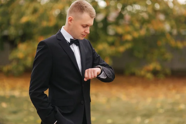 Schöner Mann im Anzug schaut draußen auf die Uhr, Herbst. Mann im schwarzen Anzug mit schöner Krawattenfliege — Stockfoto