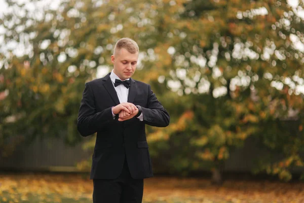 Hombre guapo de traje mira el reloj afuera, otoño. Hombre en traje negro con hermoso lazo de lazo —  Fotos de Stock