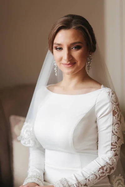 Retrato de la hermosa novia de pie junto a la ventana en casa. Encantadora novia en vestido de novia blanco —  Fotos de Stock