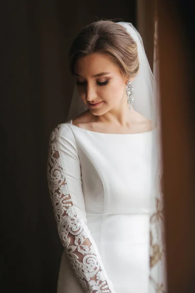 Retrato de noiva bonita em pé junto à janela em casa. Noiva encantadora em vestido de casamento branco — Fotografia de Stock