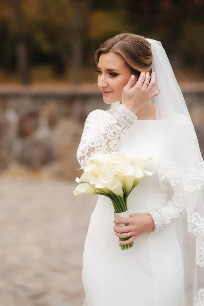 Die schöne Braut im eleganten Brautkleid steht im Park und hält einen Blumenstrauß in der Hand. Charmante Dame — Stockfoto