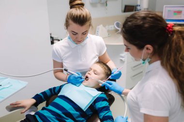 Çocuk diş hekimi ve asistan muayene erkek dişleri, o ofiste diş sandalyesinde otururken. Kadın diş hekimi